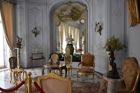 An ornate room with luxurious vintage furniture, intricate molding on the walls, and classical sculptures. The color palette includes soft whites, golds, and browns with elegant chandeliers contributing to the space. Large windows with flowing curtains allow natural light to enter, enhancing the opulence of the room. Decorative wall-mounted lights, a round table with vases, and a plush rug complete the rich interior decor.