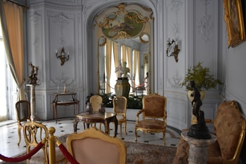 An ornate room with luxurious vintage furniture, intricate molding on the walls, and classical sculptures. The color palette includes soft whites, golds, and browns with elegant chandeliers contributing to the space. Large windows with flowing curtains allow natural light to enter, enhancing the opulence of the room. Decorative wall-mounted lights, a round table with vases, and a plush rug complete the rich interior decor.