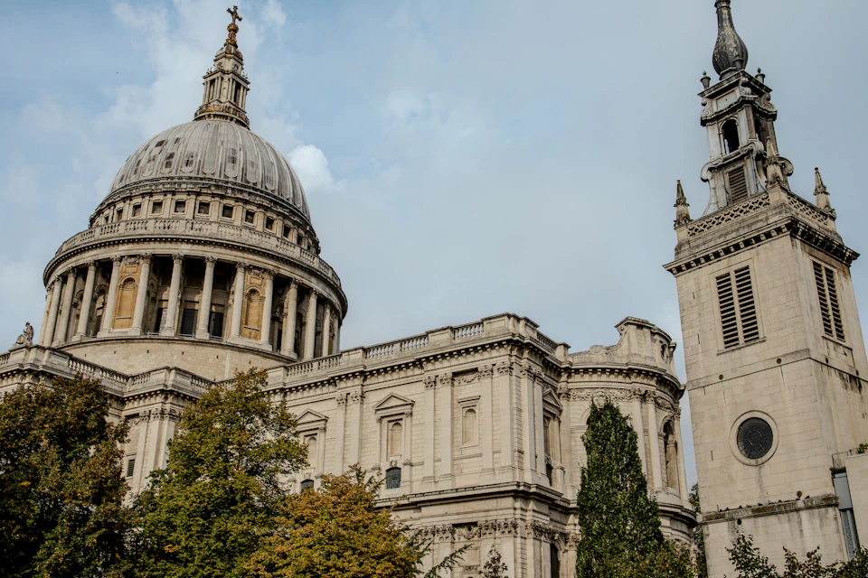 The image depicts a majestic scene featuring two prominent landmarks - the iconic dome and tower of St. Paul's Cathedral, commonly known as Big Ben, located in London, England. The cathedral is constructed from white stone and stands tall against an overcast sky with scattered clouds. The perspective of the photo is taken from below, looking up at the towering structure, emphasizing its grandeur and historical significance. In the foreground, there are trees that add a touch of nature to the urban landscape.