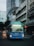 A blue city bus is driving down an urban street lined with tall buildings. The bus displays route number 53 and is illuminated against the backdrop of modern and older architecture. The street is bustling with other vehicles and motorbikes.