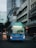 A blue city bus is driving down an urban street lined with tall buildings. The bus displays route number 53 and is illuminated against the backdrop of modern and older architecture. The street is bustling with other vehicles and motorbikes.