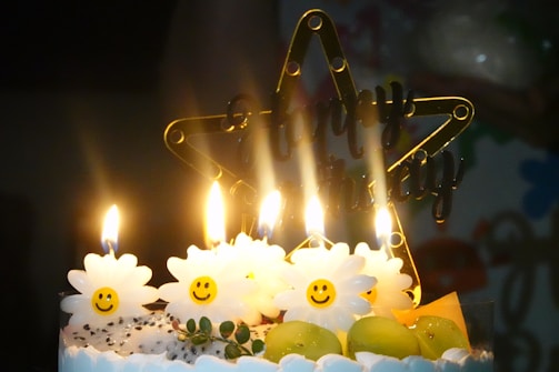 A birthday cake decorated with lit candles shaped like smiling daisies. The cake is topped with green grapes and has a decorative star with the words 'Happy Birthday'. A warm, festive glow is created by the candlelight.