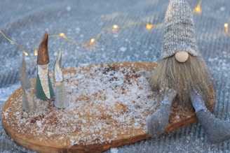 Small gnome figurines with tall hats are arranged on a wooden board dusted with artificial snow. A larger gnome with a knit hat and long beard sits to the side. Soft, glowing fairy lights are visible in the background.