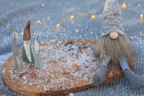 Small gnome figurines with tall hats are arranged on a wooden board dusted with artificial snow. A larger gnome with a knit hat and long beard sits to the side. Soft, glowing fairy lights are visible in the background.