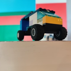 A toy vehicle constructed from colorful interlocking plastic bricks is positioned on a flat surface. The vehicle features large, rugged black wheels and a blocky body with sections in yellow, blue, and white. In the background, there is an abstract design composed of vibrant squares of red, green, and blue.