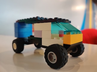 A small toy vehicle made from colorful interlocking plastic bricks sits on a smooth surface. The vehicle has four wheels and a roof made of translucent blue bricks. The body of the vehicle is composed of black, yellow, white, and blue bricks.