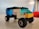 A small toy vehicle made from colorful interlocking plastic bricks sits on a smooth surface. The vehicle has four wheels and a roof made of translucent blue bricks. The body of the vehicle is composed of black, yellow, white, and blue bricks.