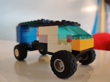 A small toy vehicle made from colorful interlocking plastic bricks sits on a smooth surface. The vehicle has four wheels and a roof made of translucent blue bricks. The body of the vehicle is composed of black, yellow, white, and blue bricks.