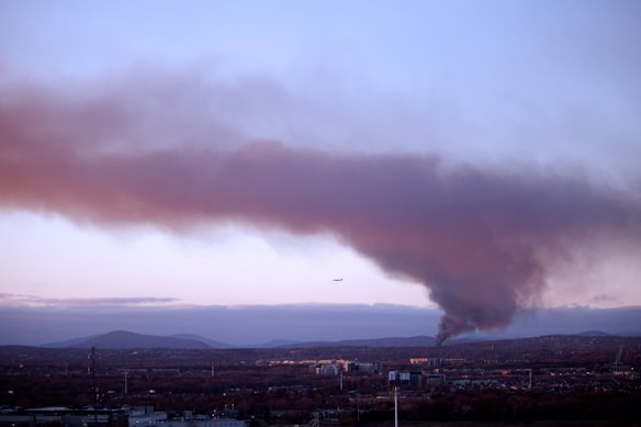 A cityscape with a large plume of dark smoke rising into the sky, indicating a fire or industrial activity. A faint silhouette of an airplane is visible in the sky near the smoke. The landscape is surrounded by distant hills.