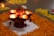 A traditional decorative arrangement featuring several oil lamps placed on a stand surrounded by bright orange marigold petals. The lamps are lit, casting a warm glow on their surroundings. Intricate designs are visible on the ground in yellow petals alongside the central floral pattern.
