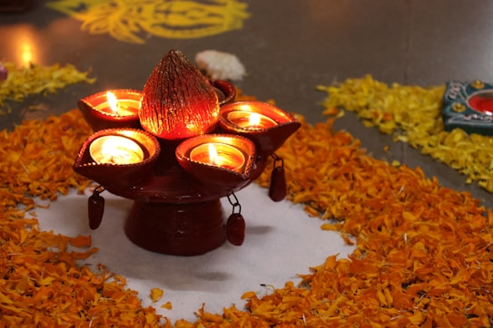 A traditional decorative arrangement featuring several oil lamps placed on a stand surrounded by bright orange marigold petals. The lamps are lit, casting a warm glow on their surroundings. Intricate designs are visible on the ground in yellow petals alongside the central floral pattern.