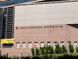A large building facade with the words 'Hospital Authority Building' and Chinese characters prominently displayed. The structure features modern architectural elements with a pattern of light brown bricks, tall glass windows on the left, and neatly trimmed greenery along the base. A yellow sign with the words 'Helping People' is visible on the left side, and a no entry sign is positioned near the middle.