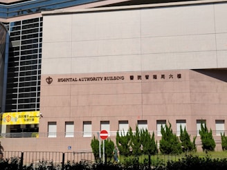 A large building facade with the words 'Hospital Authority Building' and Chinese characters prominently displayed. The structure features modern architectural elements with a pattern of light brown bricks, tall glass windows on the left, and neatly trimmed greenery along the base. A yellow sign with the words 'Helping People' is visible on the left side, and a no entry sign is positioned near the middle.