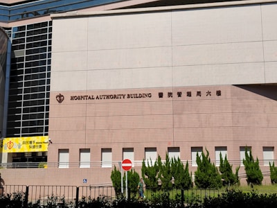 A large building facade with the words 'Hospital Authority Building' and Chinese characters prominently displayed. The structure features modern architectural elements with a pattern of light brown bricks, tall glass windows on the left, and neatly trimmed greenery along the base. A yellow sign with the words 'Helping People' is visible on the left side, and a no entry sign is positioned near the middle.