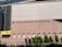 A large building facade with the words 'Hospital Authority Building' and Chinese characters prominently displayed. The structure features modern architectural elements with a pattern of light brown bricks, tall glass windows on the left, and neatly trimmed greenery along the base. A yellow sign with the words 'Helping People' is visible on the left side, and a no entry sign is positioned near the middle.