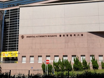 A large building facade with the words 'Hospital Authority Building' and Chinese characters prominently displayed. The structure features modern architectural elements with a pattern of light brown bricks, tall glass windows on the left, and neatly trimmed greenery along the base. A yellow sign with the words 'Helping People' is visible on the left side, and a no entry sign is positioned near the middle.