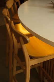 A close-up view of a modern dining area featuring light wooden chairs with curved backs and yellow seats. The chairs are arranged around a large, round, white tabletop that appears smooth and polished.