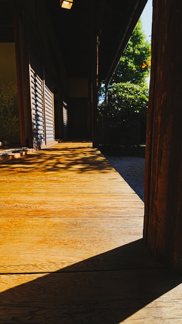 A traditional wooden deck bathed in sunlight, casting distinct shadows. The structure features wooden slats and is adjacent to a serene garden with greenery visible in the background. The warm tones of the wood contrast with the cool shadows, creating a tranquil and inviting atmosphere.