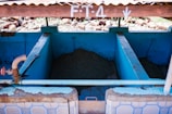 A series of concrete tanks with blue interiors, containing what appears to be agricultural material. A rusted brown pipe with a red valve is connected to the left tank. The tanks are sheltered under a corrugated metal roof with white markings, and the area outside the tanks is rocky.
