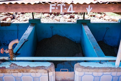 A series of concrete tanks with blue interiors, containing what appears to be agricultural material. A rusted brown pipe with a red valve is connected to the left tank. The tanks are sheltered under a corrugated metal roof with white markings, and the area outside the tanks is rocky.