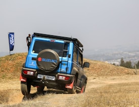 A blue Suzuki SUV is driving off-road on a grassy hill. The vehicle has a spare tire on the back with the Suzuki logo. A person stands next to a Ford flag in the distance.