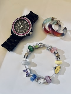 A stylish black wristwatch with a purple gemstone-studded bezel, a multicolored pair of hoop earrings with shades of blue and red, and a charm bracelet featuring various colorful charms including characters and abstract designs.