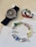 A stylish black wristwatch with a purple gemstone-studded bezel, a multicolored pair of hoop earrings with shades of blue and red, and a charm bracelet featuring various colorful charms including characters and abstract designs.