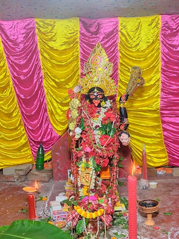 A richly adorned statue of a deity, surrounded by vibrant garlands and traditional ornaments, placed against a backdrop of alternating pink and yellow draped fabric. The setting includes burning candles and incense, with various ceremonial items like a banana leaf, a green bottle, and small bowls scattered around the base.