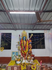A detailed and colorful altar dedicated to a Hindu goddess features an intricately decorated idol adorned with vibrant garlands and offerings. The setting includes a variety of flowers, fruits, and other religious items arranged meticulously. The space is illuminated with twinkling lights and is in a room with a corrugated metal ceiling and a ceiling fan. Windows with black grills are seen in the background.