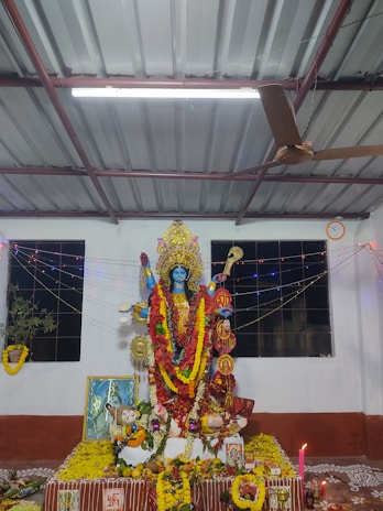 A detailed and colorful altar dedicated to a Hindu goddess features an intricately decorated idol adorned with vibrant garlands and offerings. The setting includes a variety of flowers, fruits, and other religious items arranged meticulously. The space is illuminated with twinkling lights and is in a room with a corrugated metal ceiling and a ceiling fan. Windows with black grills are seen in the background.