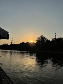 The image captures a sunset over a river, with a silhouette of trees and buildings against the sky. The sun is partially hidden, creating a golden glow on the water surface.
