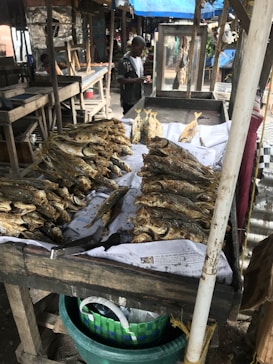 A market stall displaying numerous dried fish arranged neatly on a table covered with newspaper. The setting appears to be an outdoor market with wooden stalls and a person in the background looking at a phone.
