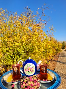 A vibrant outdoor scene with a tea set placed on a tray. The set includes a blue teapot with floral designs, two glasses of tea, and bowls containing sugar cubes and dried rosebuds. In the background, yellow and green foliage contrasts with a clear blue sky.