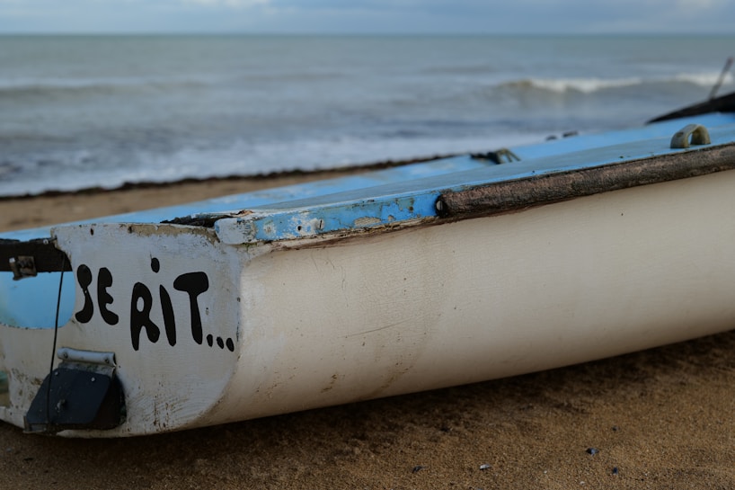 A weathered boat rests on a sandy beach, with a part of the ocean visible in the background. The side of the boat has the faded text 'SE RIT...' painted in bold black letters. The sky is cloudy, hinting at a calm, overcast day.