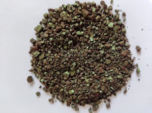 A collection of small, irregular, metallic-colored granules scattered on a white surface. The granules have a shiny, reflective surface with a greenish tint.