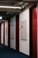 A corridor with blue carpet flooring, red and white walls, and bright overhead lighting. Bulletin boards are mounted on the walls, some with posted notices.