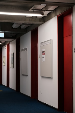 A corridor with blue carpet flooring, red and white walls, and bright overhead lighting. Bulletin boards are mounted on the walls, some with posted notices.
