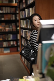 A woman with a striped black and white shirt stands in a bookstore, carrying a large black handbag. She appears to be browsing through the shelves filled with books. The shelves are lined with various colorful book spines.
