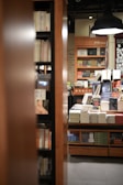A cozy bookstore setting featuring shelves filled with books. There is a stack of neatly organized books on a central wooden table, partially obscured by bookshelves in the foreground. The warm lighting and wooden furnishings contribute to an inviting and intimate atmosphere.