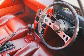 A vintage car interior featuring a red leather upholstery. The dashboard includes a classic analog speedometer and other gauges. A black steering wheel with metal accents and a gear shift is prominently visible. The overall design gives a retro automotive vibe.