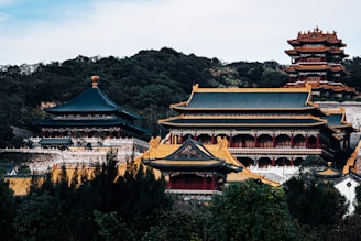 A traditional Asian palace complex with ornate roofing and multiple tiers stands amidst lush greenery. The architecture features intricate details, with vibrant colors and decorative elements.