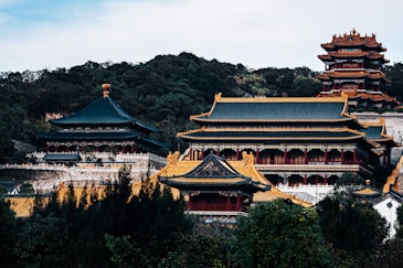 A traditional Asian palace complex with ornate roofing and multiple tiers stands amidst lush greenery. The architecture features intricate details, with vibrant colors and decorative elements.