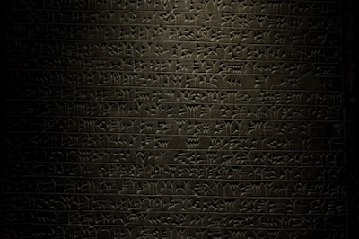 A stone tablet displaying rows of ancient cuneiform script with intricate symbols carved into its surface. The text is written in a uniform pattern, and the carvings are highlighted by subtle lighting, emphasizing the ancient craftsmanship.