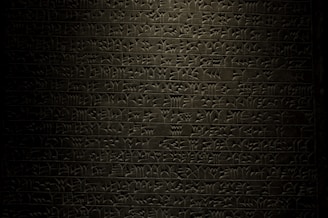 A stone tablet displaying rows of ancient cuneiform script with intricate symbols carved into its surface. The text is written in a uniform pattern, and the carvings are highlighted by subtle lighting, emphasizing the ancient craftsmanship.