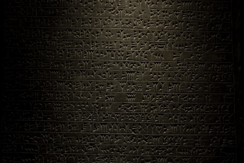 A stone tablet displaying rows of ancient cuneiform script with intricate symbols carved into its surface. The text is written in a uniform pattern, and the carvings are highlighted by subtle lighting, emphasizing the ancient craftsmanship.