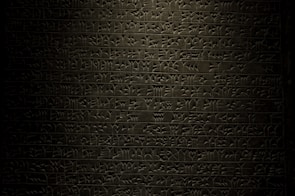 A stone tablet displaying rows of ancient cuneiform script with intricate symbols carved into its surface. The text is written in a uniform pattern, and the carvings are highlighted by subtle lighting, emphasizing the ancient craftsmanship.
