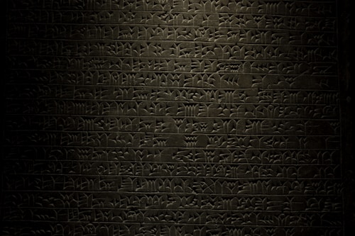 A stone tablet displaying rows of ancient cuneiform script with intricate symbols carved into its surface. The text is written in a uniform pattern, and the carvings are highlighted by subtle lighting, emphasizing the ancient craftsmanship.