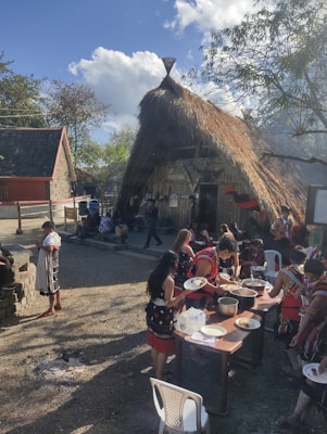 A group of people are gathered outside near a thatched roof structure, engaging in a communal meal. They are wearing traditional attire, and serving food from a table set with pots and plates. The setting is outdoors, with clear blue skies and some trees in the background.