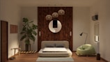 A modern living room features a low-profile sofa bed in the center, set against a wooden panel wall with a round decorative mirror. Above the sofa hang several pendant lights. To the left, a large potted plant adds a touch of greenery, next to a small wooden stool. On the right, a stylish floor lamp stands beside a green, plush armchair. The room also has an air conditioning unit and a window with a rolled-up blind.