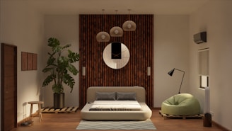 A modern living room features a low-profile sofa bed in the center, set against a wooden panel wall with a round decorative mirror. Above the sofa hang several pendant lights. To the left, a large potted plant adds a touch of greenery, next to a small wooden stool. On the right, a stylish floor lamp stands beside a green, plush armchair. The room also has an air conditioning unit and a window with a rolled-up blind.
